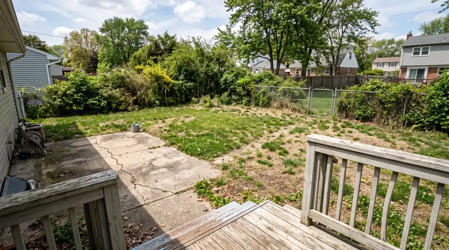 The yard before: a patchy lawn with bare dirt, an overgrown shrub border, a cracked concrete slab and a chain-link fence.